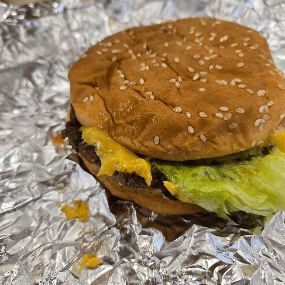 A sesame seed hamburger bun filled with a beef patty, melted cheese, and lettuce, wrapped in foil.